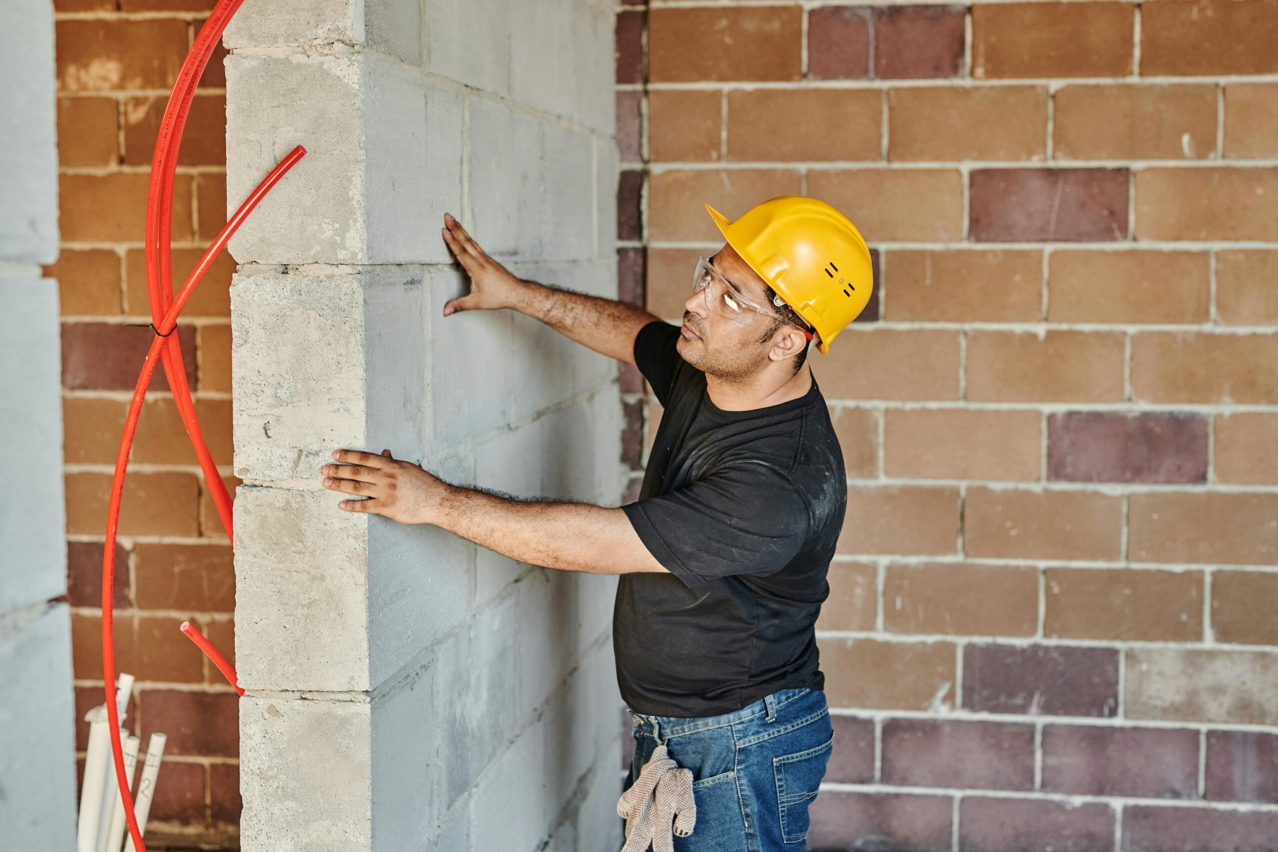pexels photo 8961703 8961703 Man in hard hat checks brick wall in indoor construction site.
